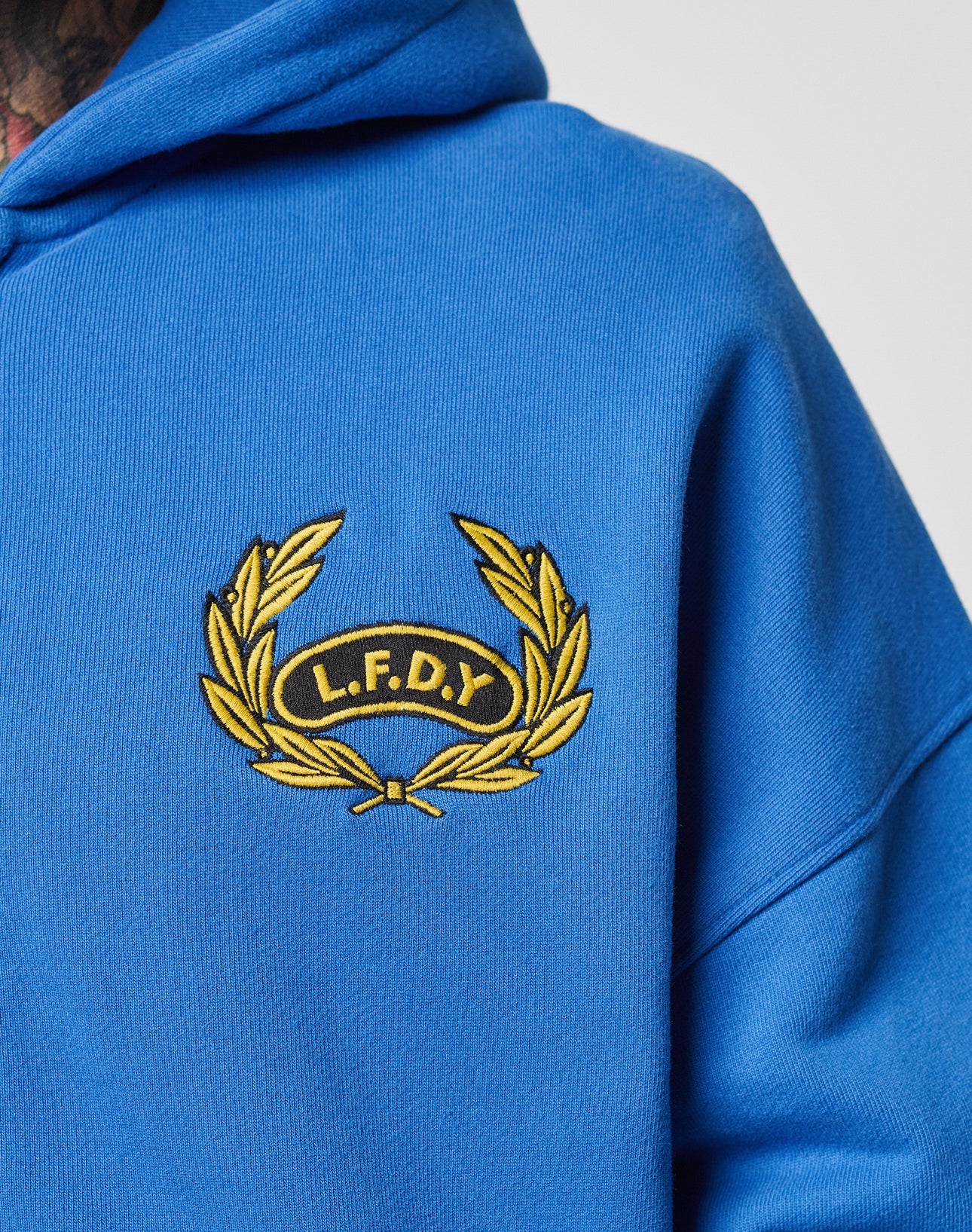 A close-up of the LFDY Team Hooded Zip by LIVE FAST DIE YOUNG in Oversize Fit, showcasing an embroidered yellow laurel wreath with the initials L.F.D.Y on the left chest.