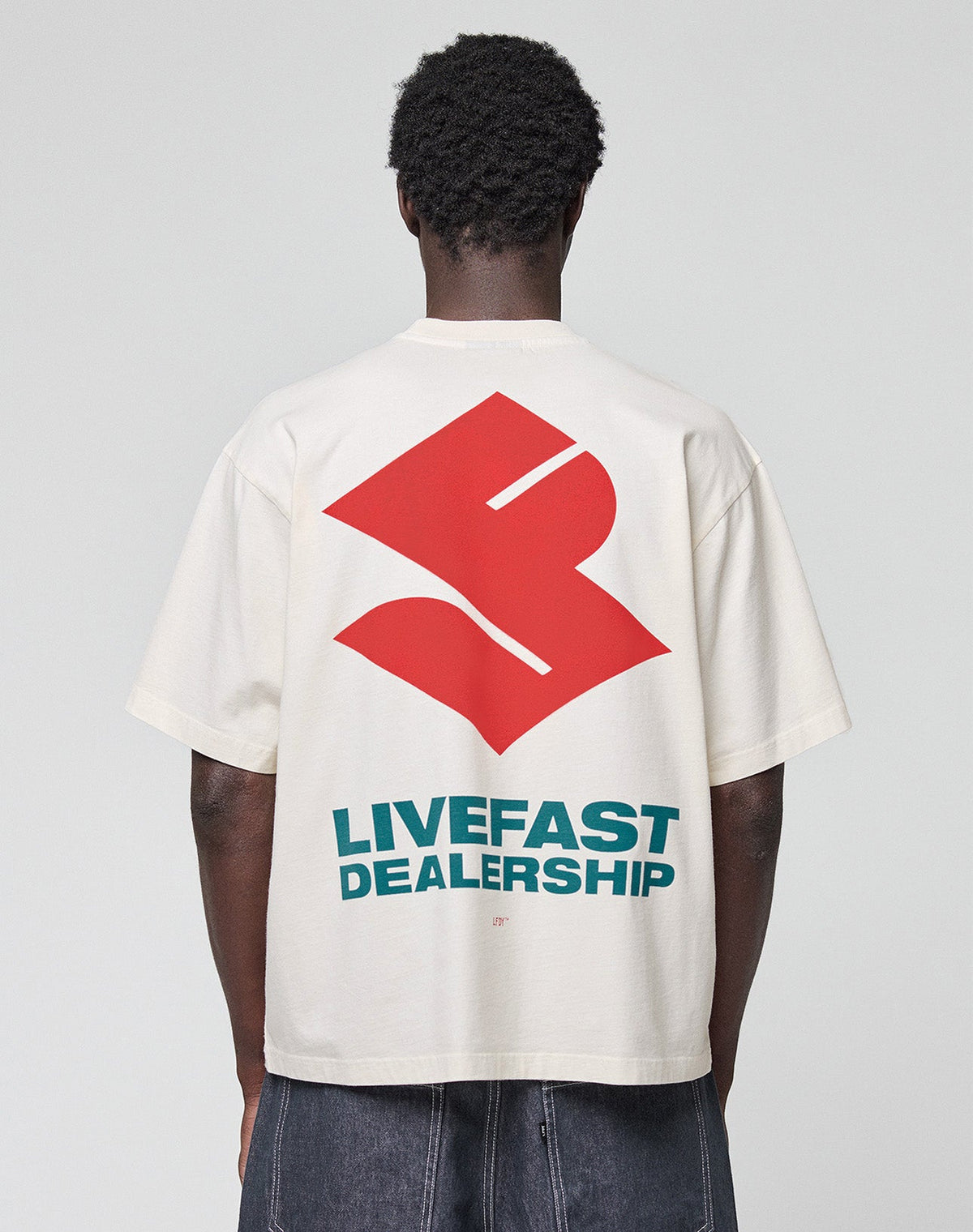 A person faces away, wearing the LFDY Dealership Tee by LIVE FAST DIE YOUNG—an oversize white 100% Baumwolle shirt with a bold red abstract backprint and "LIVEFAST DEALERSHIP" in striking teal letters. | model