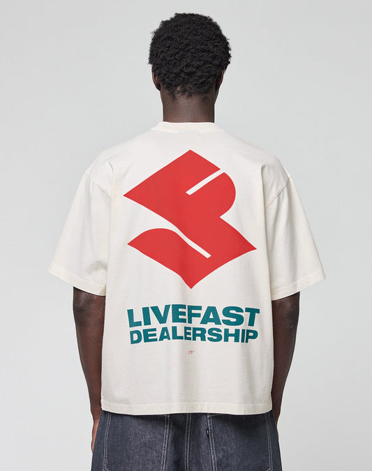 A person faces away, wearing the LFDY Dealership Tee by LIVE FAST DIE YOUNG—an oversize white 100% Baumwolle shirt with a bold red abstract backprint and "LIVEFAST DEALERSHIP" in striking teal letters. | model