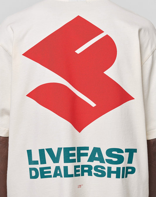 A person wears the LFDY Dealership Tee by LIVE FAST DIE YOUNG, an oversized white 100% Baumwolle shirt with a bold red abstract back graphic and teal "LIVEFAST DEALERSHIP" text, featuring striking front and back prints.