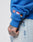 A person wears the LFDY Team Hooded Zip by LIVE FAST DIE YOUNG—an oversize blue sweatshirt with UK, Dutch, and German flag embroidery on the sleeve. They have a tattooed hand and pair it with light-washed denim jeans.