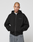 A person with a shaved head wears the Basic 510 Hooded Zip by LIVE FAST DIE YOUNG in black Oversize Fit, with LiveFast text and front/back prints, paired with gray jeans. They stand hands-in-pockets against a plain light gray background.
