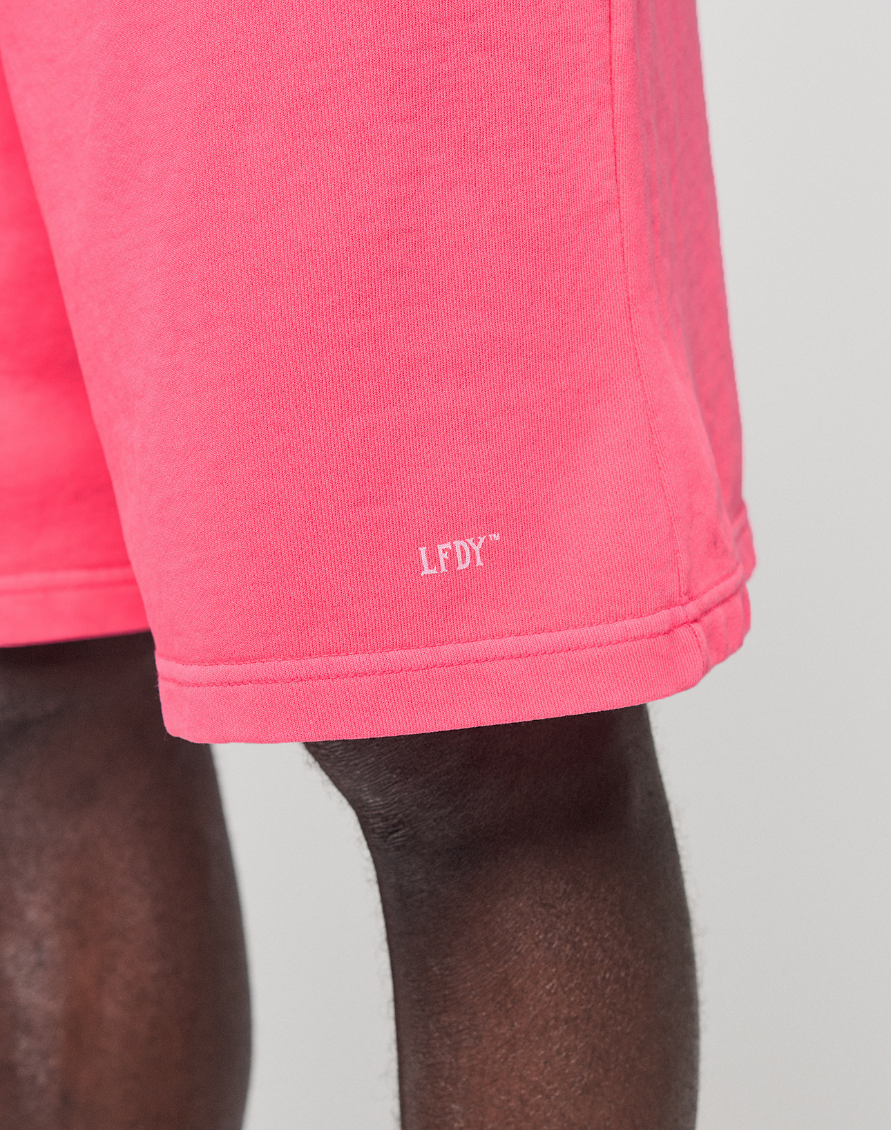 Close-up of a person wearing bright pink Basic Shorts by LIVE FAST DIE YOUNG, featuring an elastic waistband and white LFDY™ logo print near the hem, set against a plain neutral background.