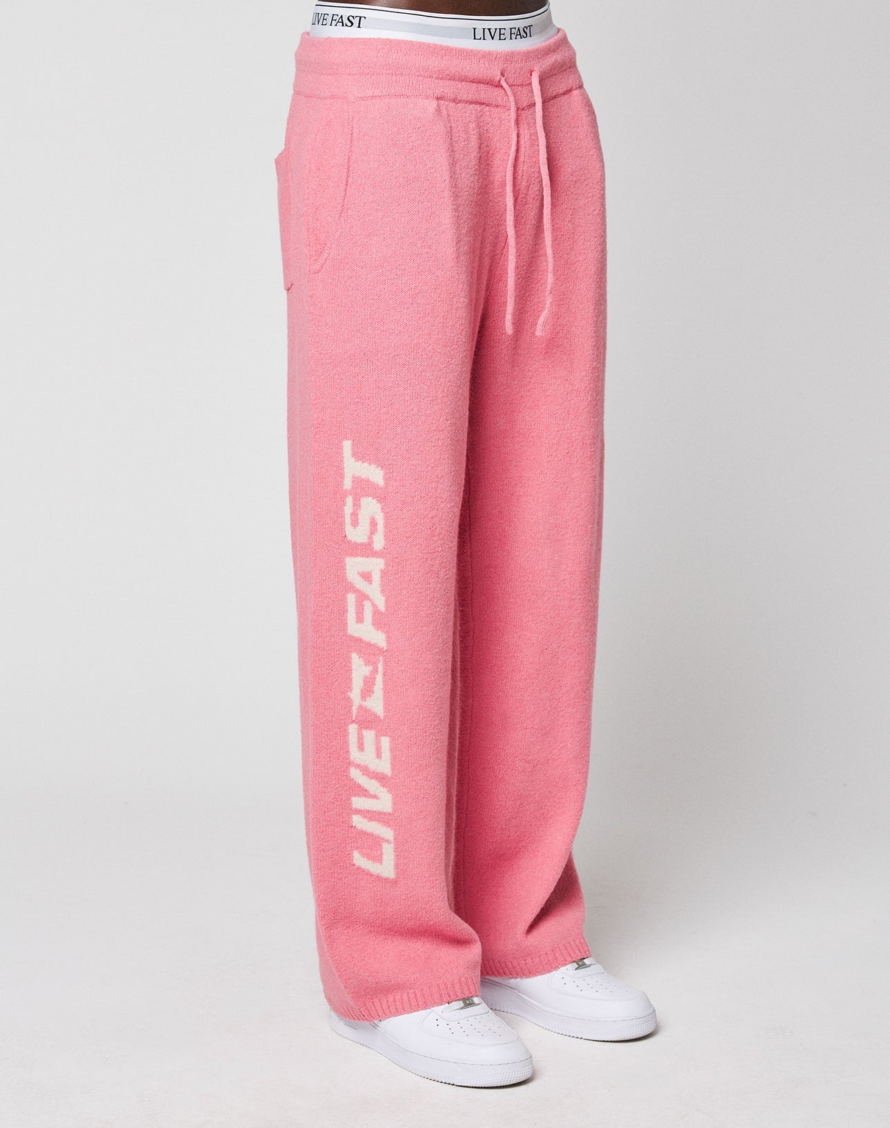 A person wears LIVE FAST DIE YOUNG’s Live Fast Boucle Pants—loose-fitting, wide-leg, pink with “LIVE FAST” in white on the left leg and elastic waistband—paired with white sneakers.