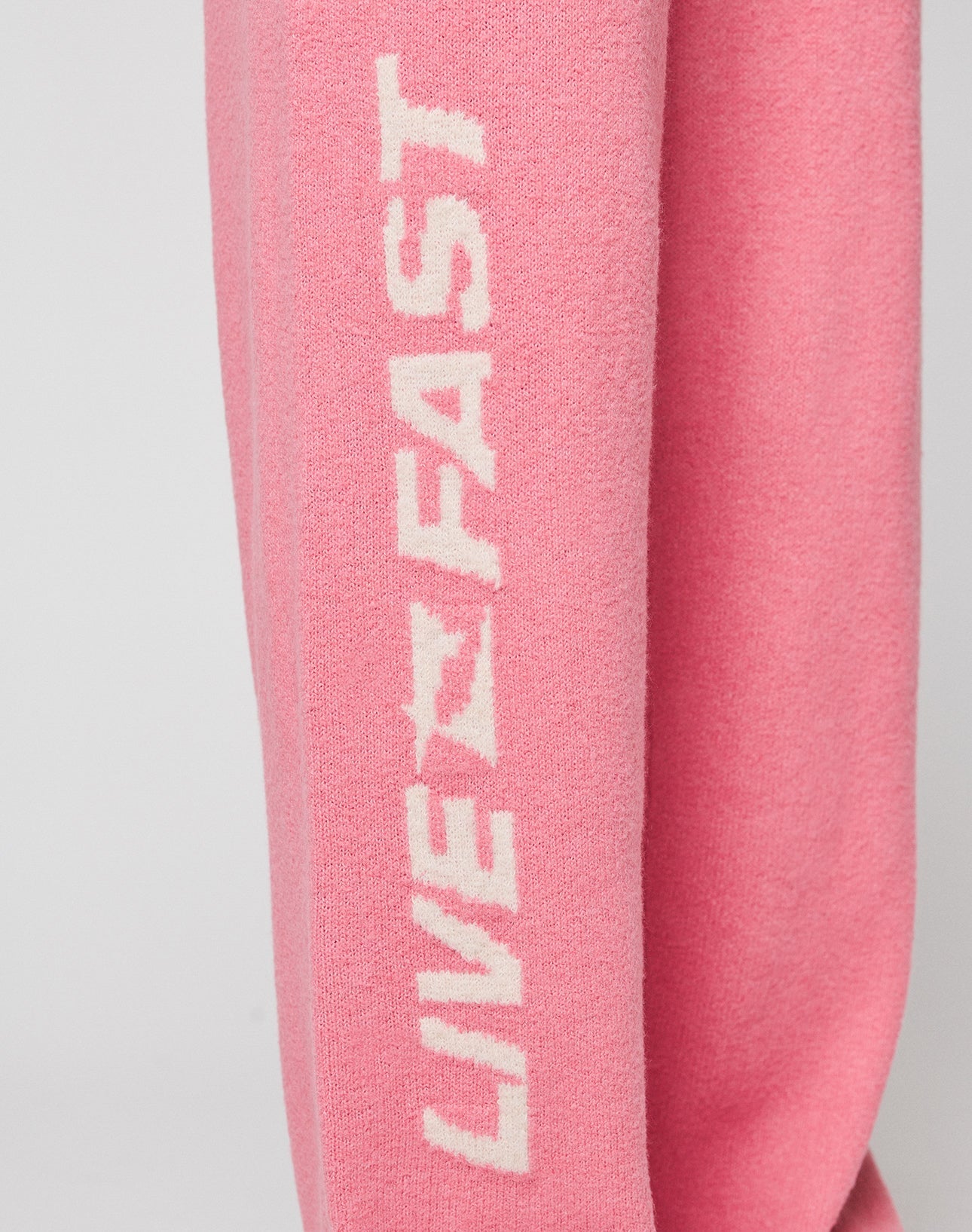 Close-up of pink fabric on the Live Fast Boucle Pants by LIVE FAST DIE YOUNG, with bold white "LIVE FAST" lettering printed vertically along the leg, featuring a straight leg (gerades Bein) and elastic waistband.