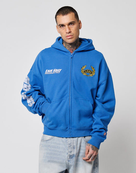 A man with short hair and neck tattoos wears the LIVE FAST DIE YOUNG LFDY Team Hooded Zip in an oversize fit, featuring multiple logos, embroidery, and a flag patch, paired with light blue jeans against a plain white background.