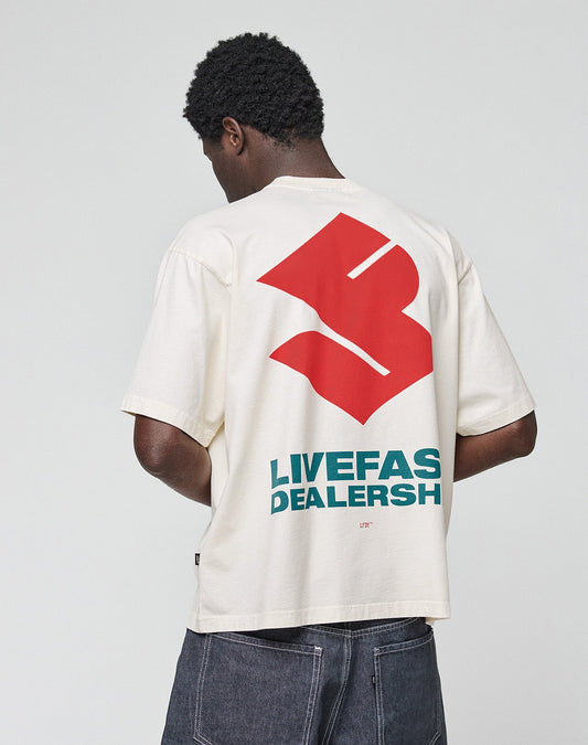 A person with short curly hair is seen from behind, wearing the cream LFDY Dealership Tee by LIVE FAST DIE YOUNG, featuring a bold red graphic and green LIVE FAS DEALERSH text on the back against a plain background.