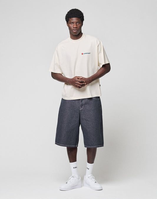 A man stands against a plain backdrop, wearing the LIVE FAST DIE YOUNG LFDY Dealership Tee in beige (100% Baumwolle), loose dark denim shorts, white socks, and white sneakers. He faces forward with his hands clasped in front.