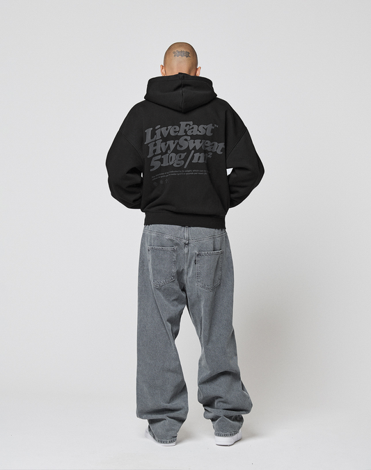 A person with a shaved head, seen from behind, wears the LIVE FAST DIE YOUNG Basic 510 Hooded Zip in black with gray text and loose-fitting gray Vintage-Waschung jeans, set against a plain white background.