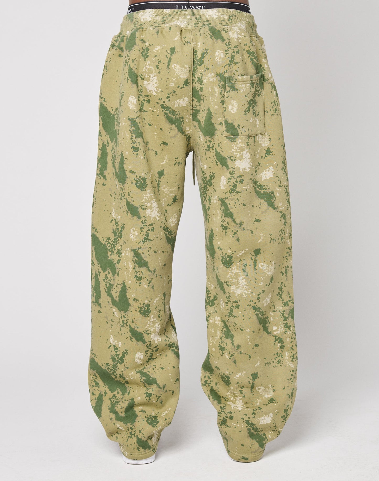 A person is seen from the back wearing LIVE FAST DIE YOUNGs Camo Wide Jogger—tan, wide-fit pants with green and white splatter camouflage and an elastic waistband—posed against a plain white background.