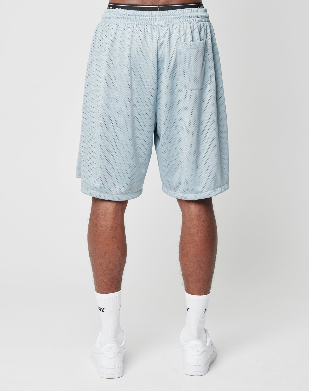 A person faces away wearing LIVE FAST DIE YOUNG LFDY Loyal Shorts in light blue mesh polyester with an elastic waistband and a small right back pocket, paired with white crew socks and sneakers, against a plain white background.