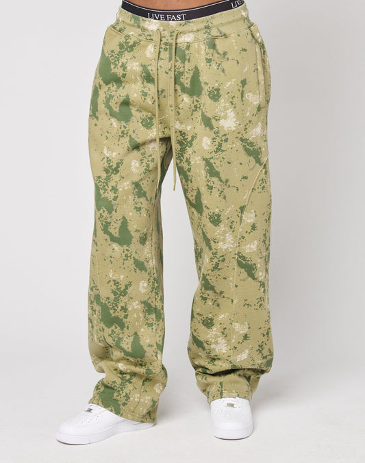 A person wearing the Camo Wide Jogger by LIVE FAST DIE YOUNG, featuring a green and beige camouflage print, wide fit, elastic waistband, and styled with white sneakers, stands against a plain white background.