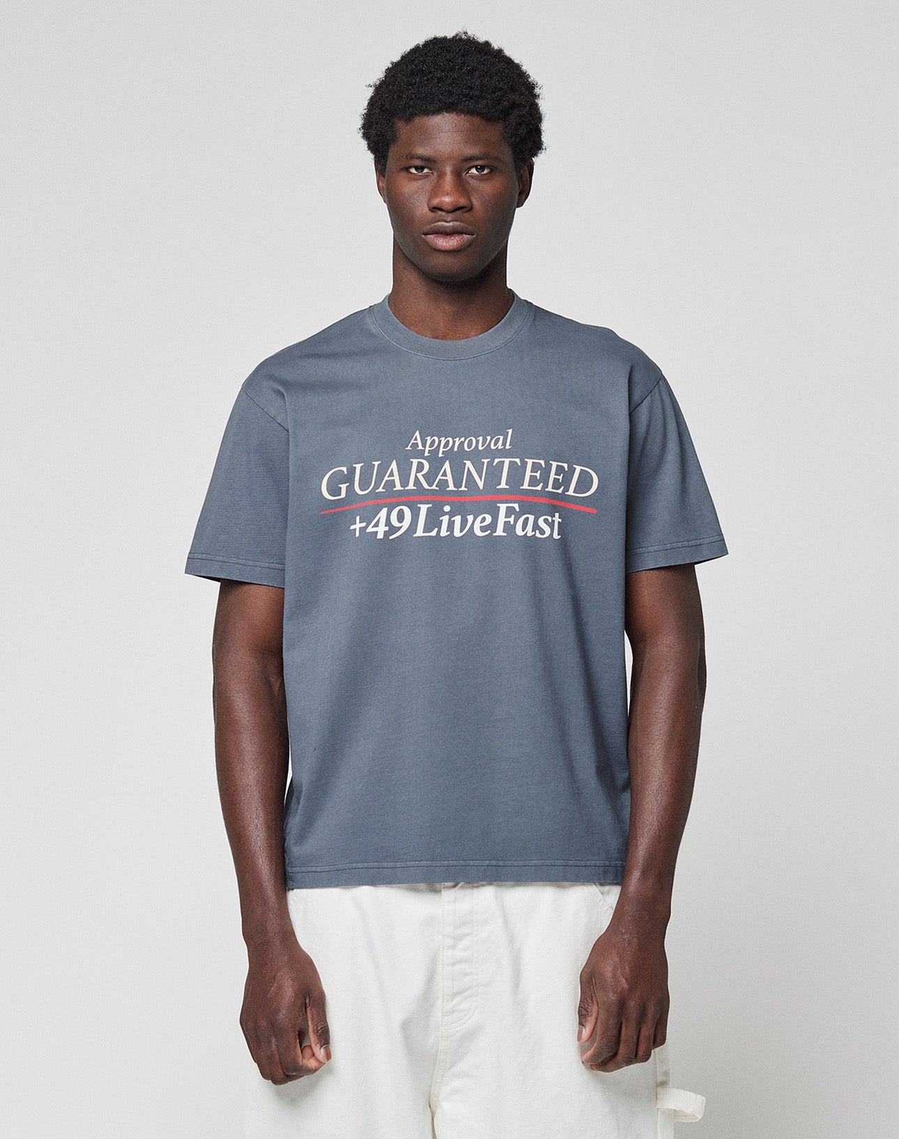 A young man wears the LFDY Guaranteed Tee by LIVE FAST DIE YOUNG, a regular fit gray t-shirt made of 100% cotton with bold “Approval GUARANTEED +49LiveFast” front print, paired with white pants against a light gray background.