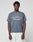 A young man wears the LFDY Guaranteed Tee by LIVE FAST DIE YOUNG, a regular fit gray t-shirt made of 100% cotton with bold “Approval GUARANTEED +49LiveFast” front print, paired with white pants against a light gray background.