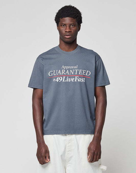 A young man wears the LFDY Guaranteed Tee by LIVE FAST DIE YOUNG, a regular fit gray t-shirt made of 100% cotton with bold “Approval GUARANTEED +49LiveFast” front print, paired with white pants against a light gray background.