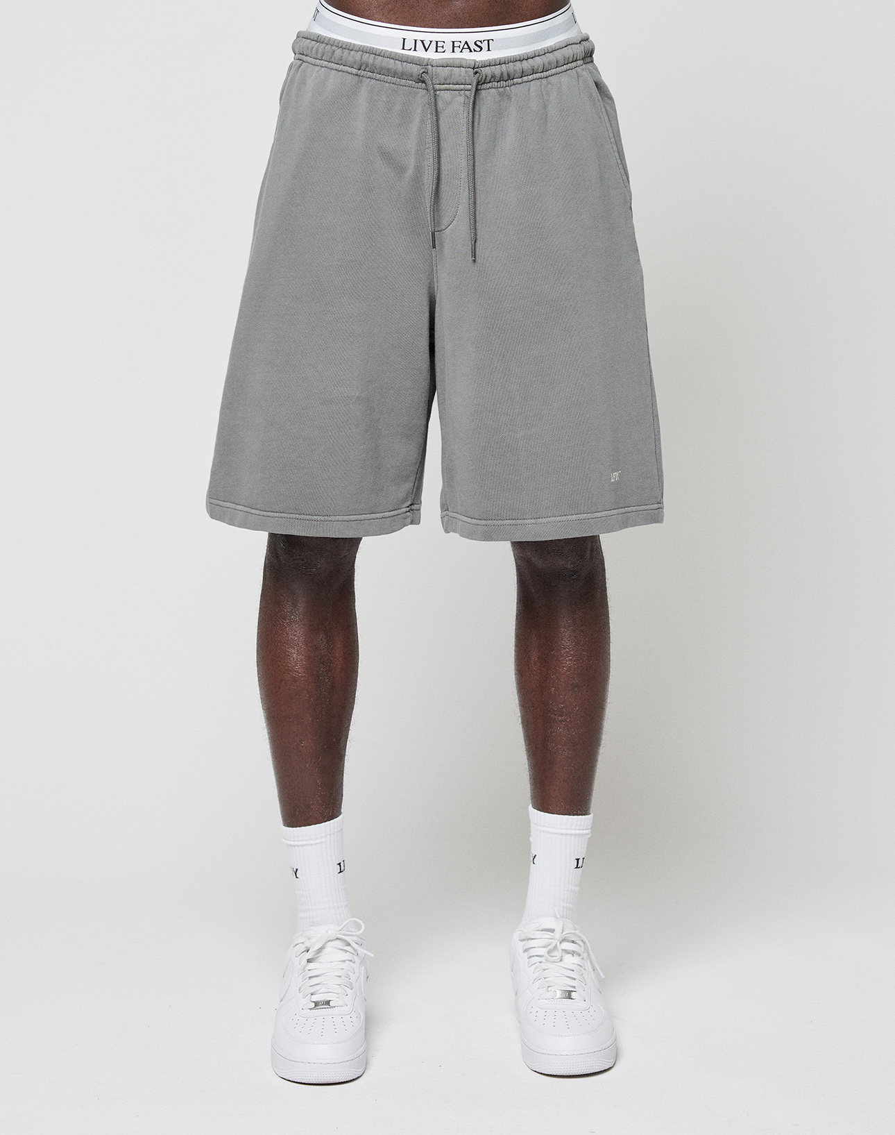 A person wears LIVE FAST DIE YOUNG Basic Shorts in regular fit gray with a faded finish and a white waistband reading "LIVE FAST," paired with white socks and sneakers, standing against a plain white background. Upper body not shown.