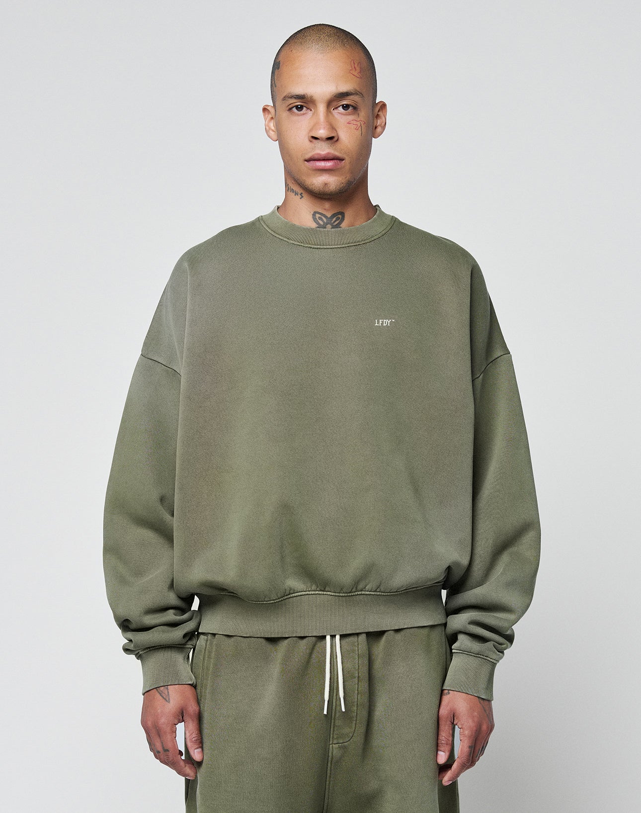 A person wears the LIVE FAST DIE YOUNG Basic Chest Sweater, an oversize fit olive green sweatshirt with subtle lettering and a handgefertigter Fade-Effekt, paired with matching drawstring sweatpants against a light gray background.