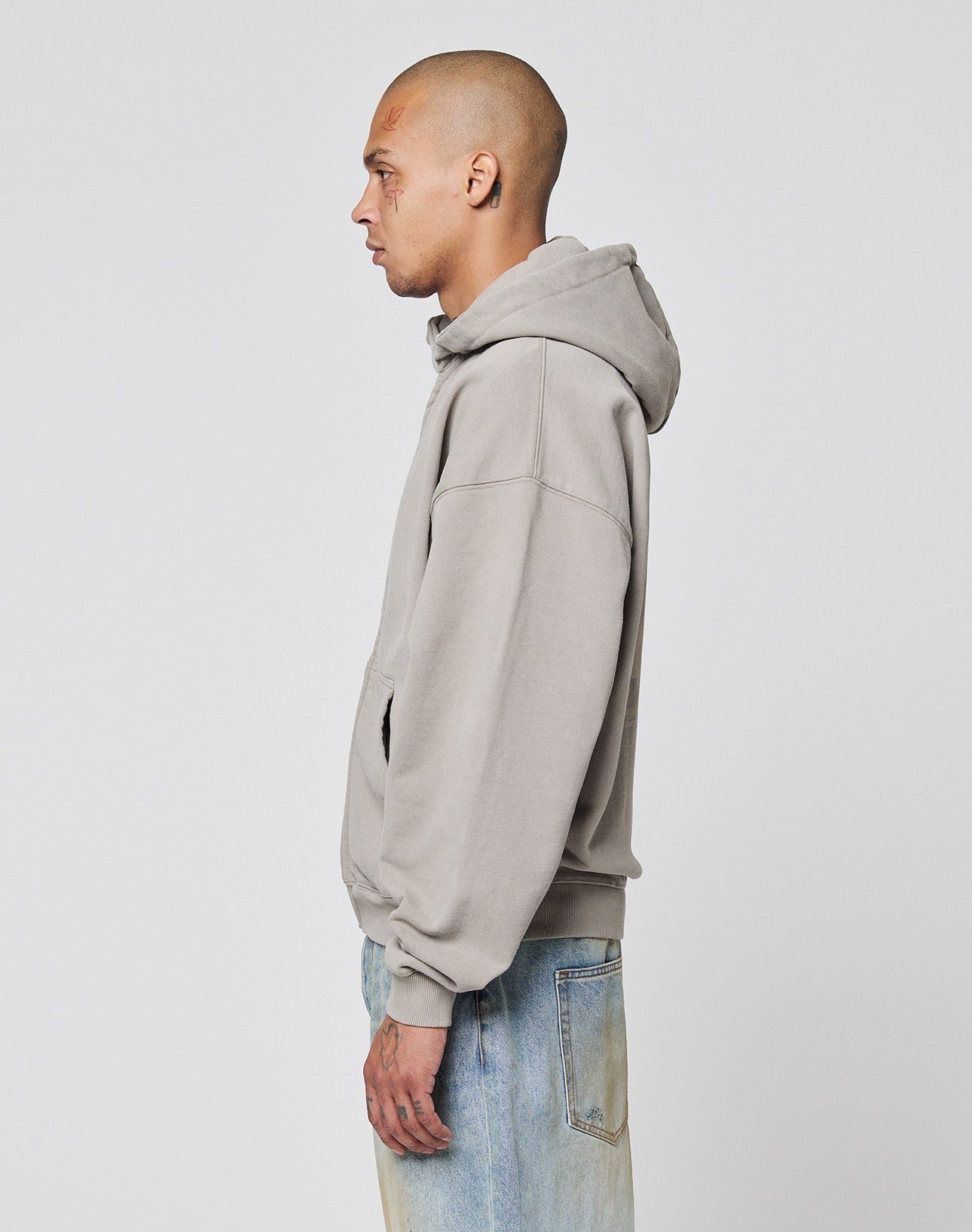 A person with a shaved head stands in profile against a plain background, wearing the LIVE FAST DIE YOUNG Basic 510 Hooded Zip hoodie in light gray and faded blue jeans in vintage wash.