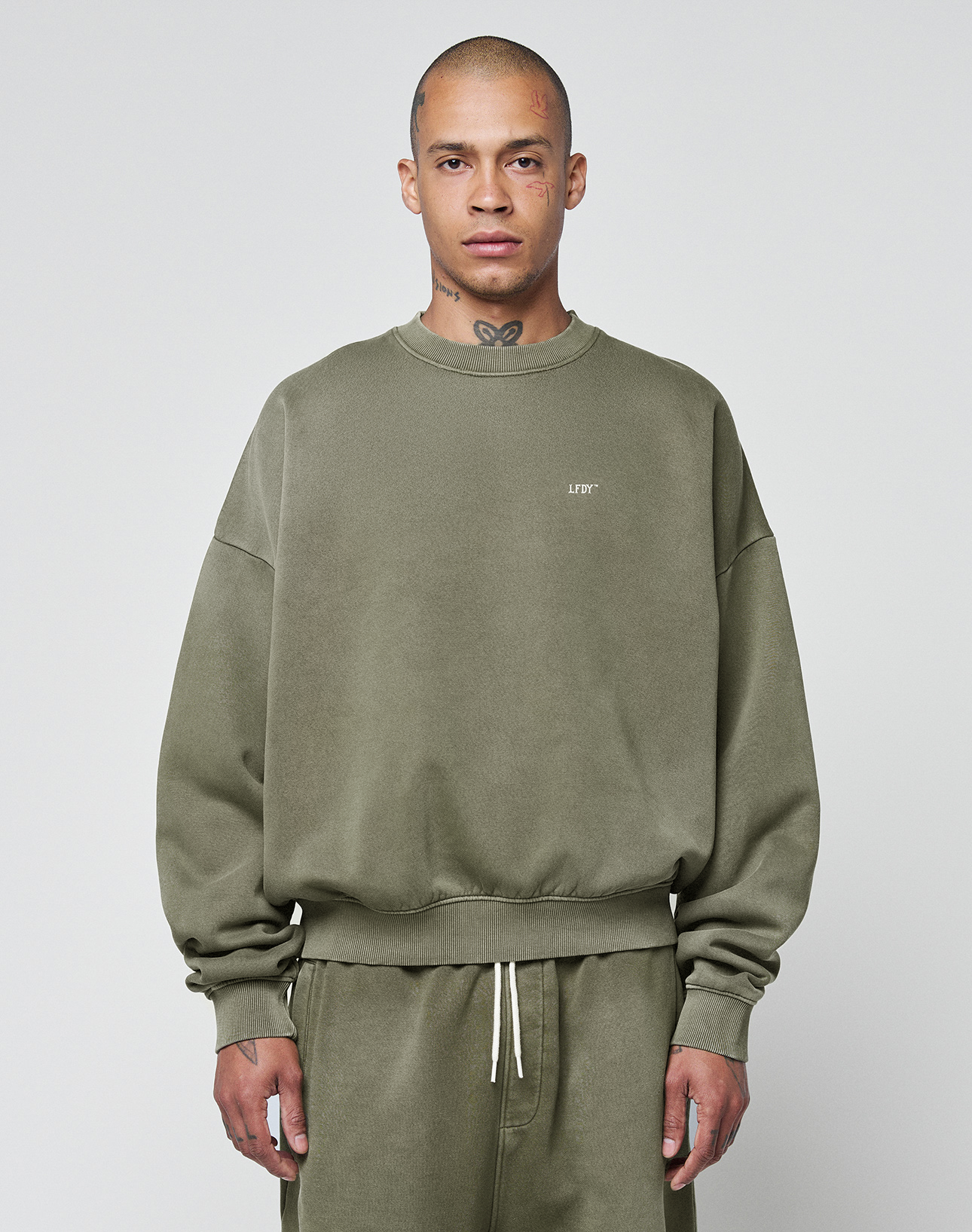 A person wears the LIVE FAST DIE YOUNG Basic Chest Sweater in olive green with a handmade fade effect and matching sweatpants, standing facing forward against a light gray background. The oversize fit sweater features small chest text. | model