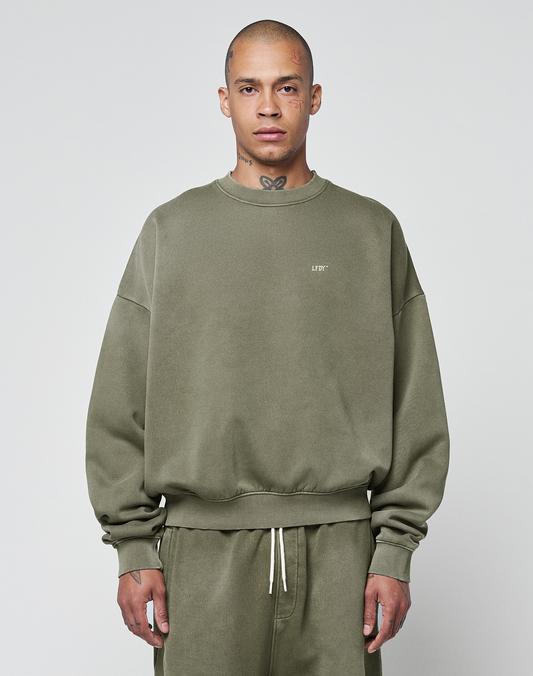 A person wears the LIVE FAST DIE YOUNG Basic Chest Sweater in olive green with a handmade fade effect and matching sweatpants, standing facing forward against a light gray background. The oversize fit sweater features small chest text. | model