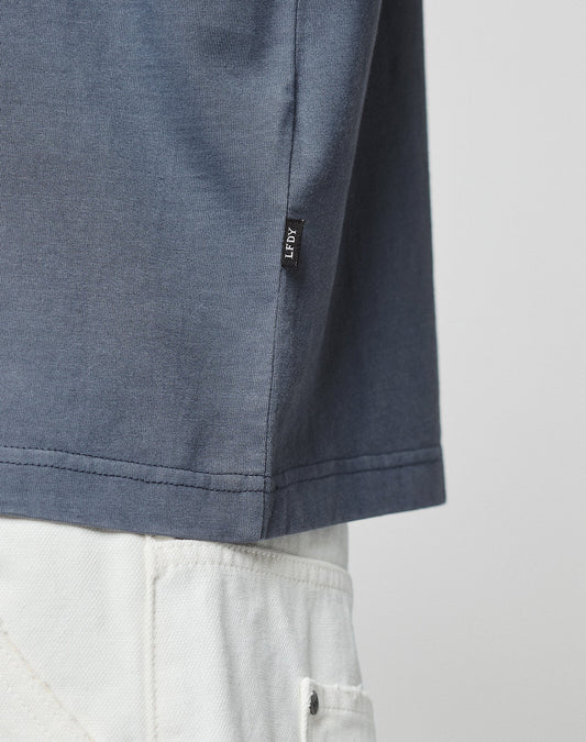 A close-up showing the hem of the LFDY Guaranteed Tee (LIVE FAST DIE YOUNG) in blue, made from 100% Baumwolle, worn with white pants. A small black LEFTY tag is visible. The background is plain and neutral.