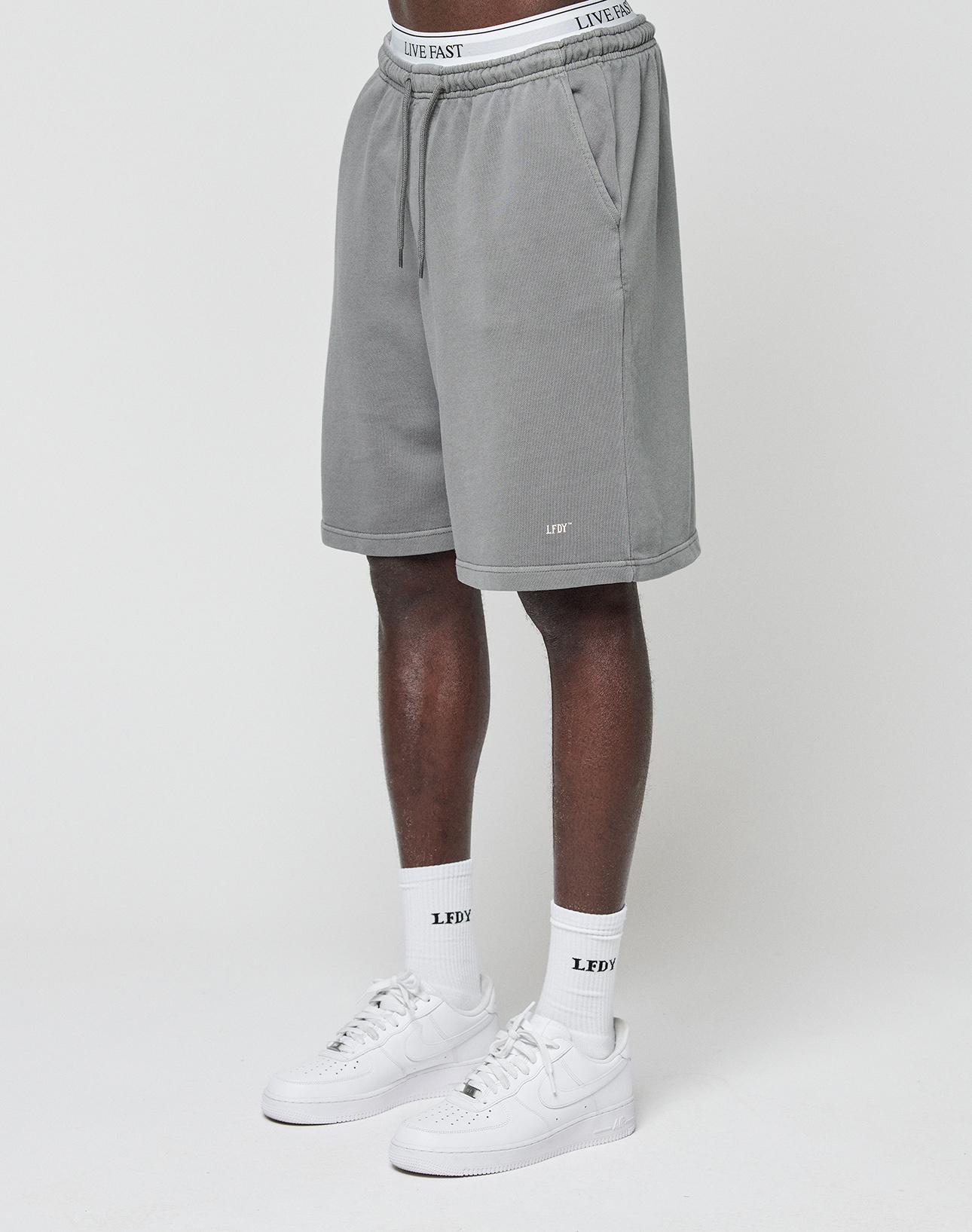 A person wears LIVE FAST DIE YOUNG Basic Shorts in gray with a branded waistband, white LEDY logo socks, and white sneakers. Only the lower half of the regular fit look is shown against a plain white background.