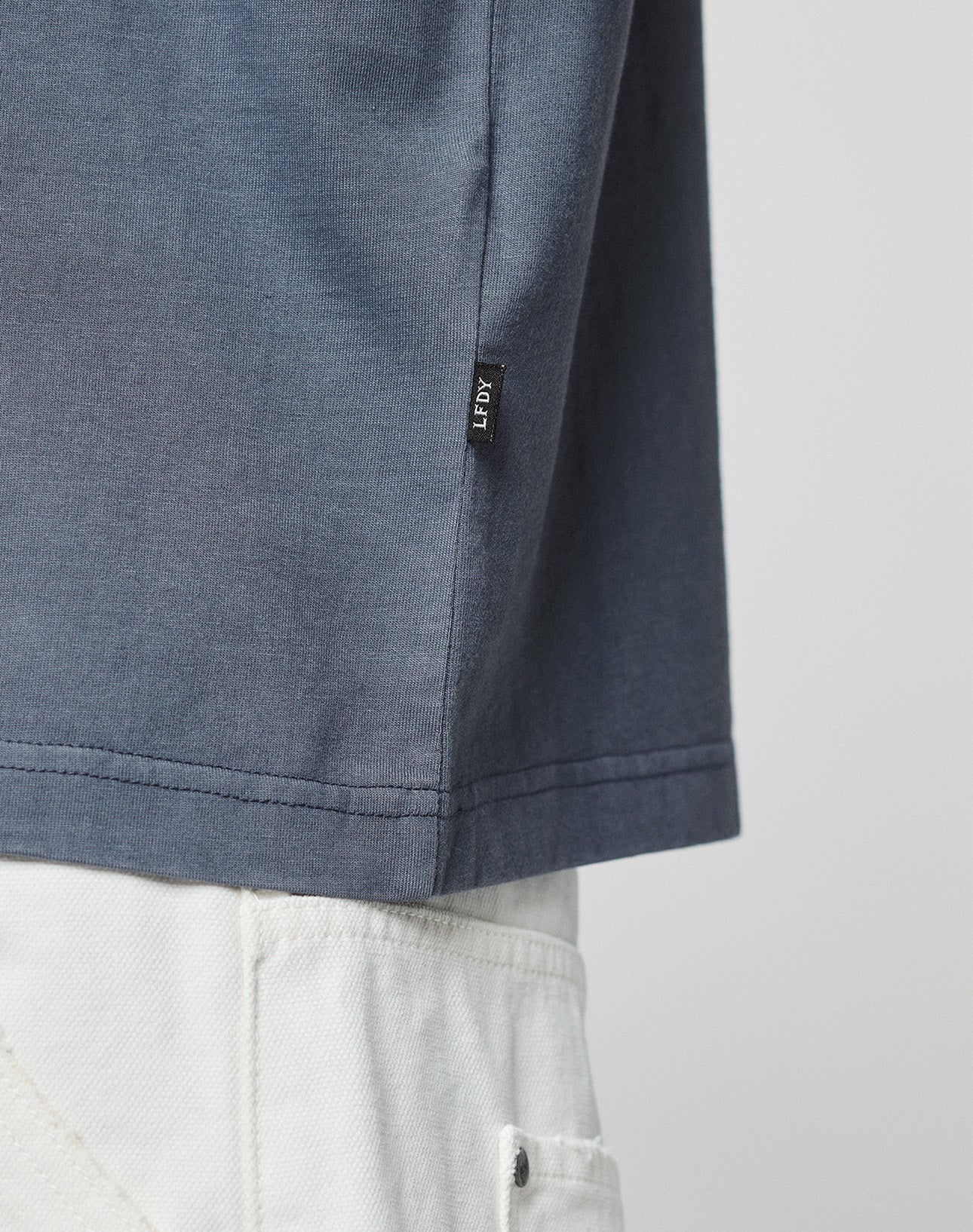 A close-up showing the hem of the LFDY Guaranteed Tee (LIVE FAST DIE YOUNG) in blue, made from 100% Baumwolle, worn with white pants. A small black LEFTY tag is visible. The background is plain and neutral.