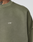 Close-up of a person wearing the LIVE FAST DIE YOUNG Basic Chest Sweater in olive green with white LFDY™ text on the chest and an Oversize Fit. The neck, part of the shoulder, and a neck tattoo are visible.