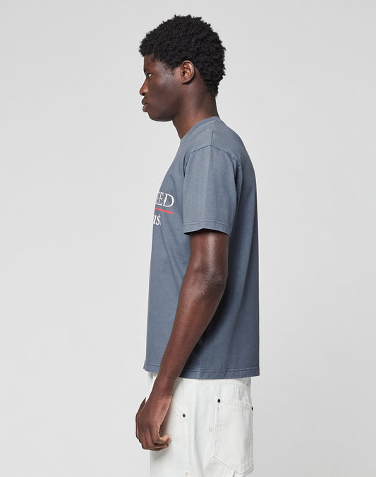 A person with short curly hair stands in profile against a plain background, wearing a gray LFDY Guaranteed Tee by LIVE FAST DIE YOUNG and light-colored pants.