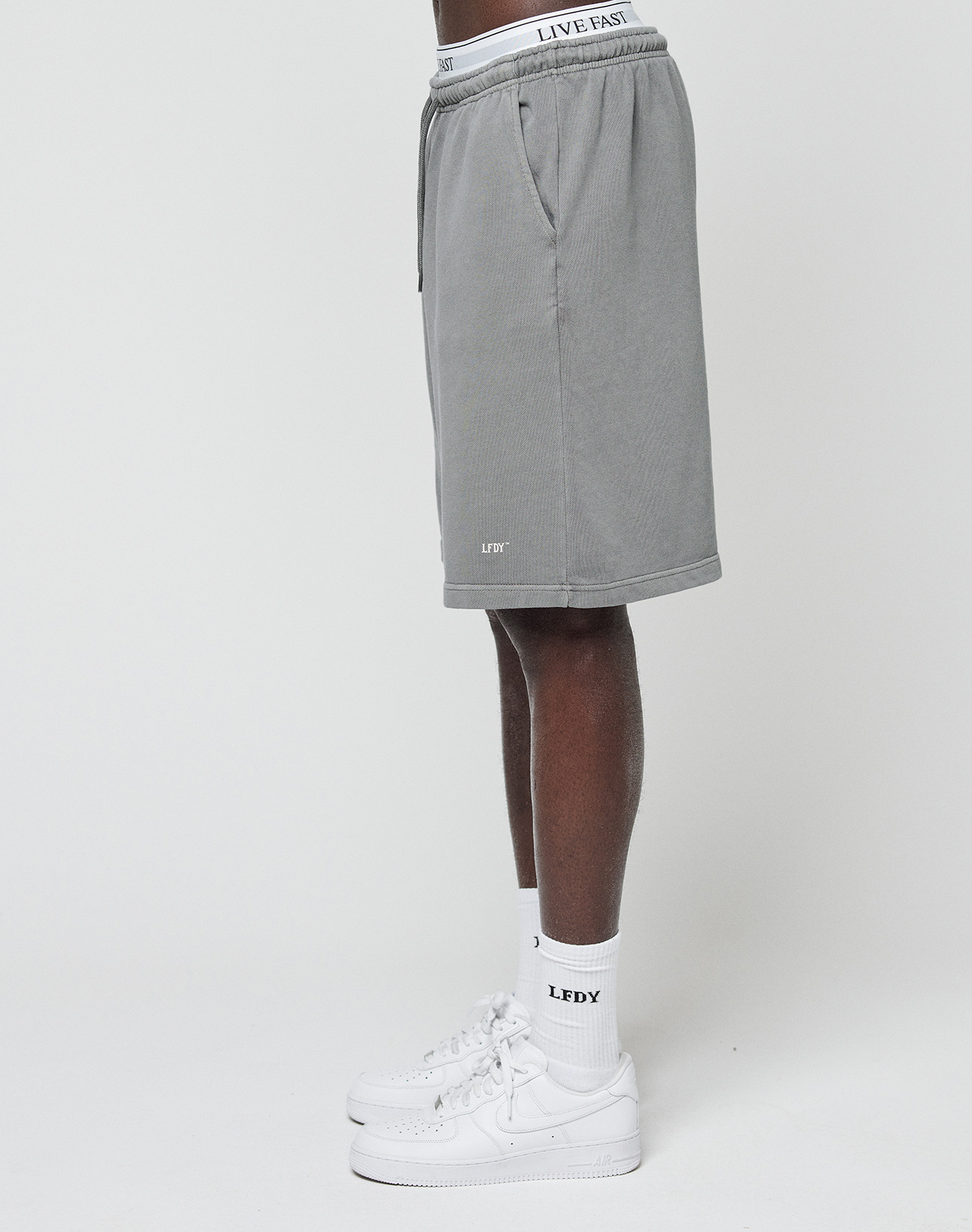 A person in LIVE FAST DIE YOUNG Basic Shorts, white LEDY logo socks, white sneakers, and underwear with a LIVE FAST waistband stands against a plain white background.
