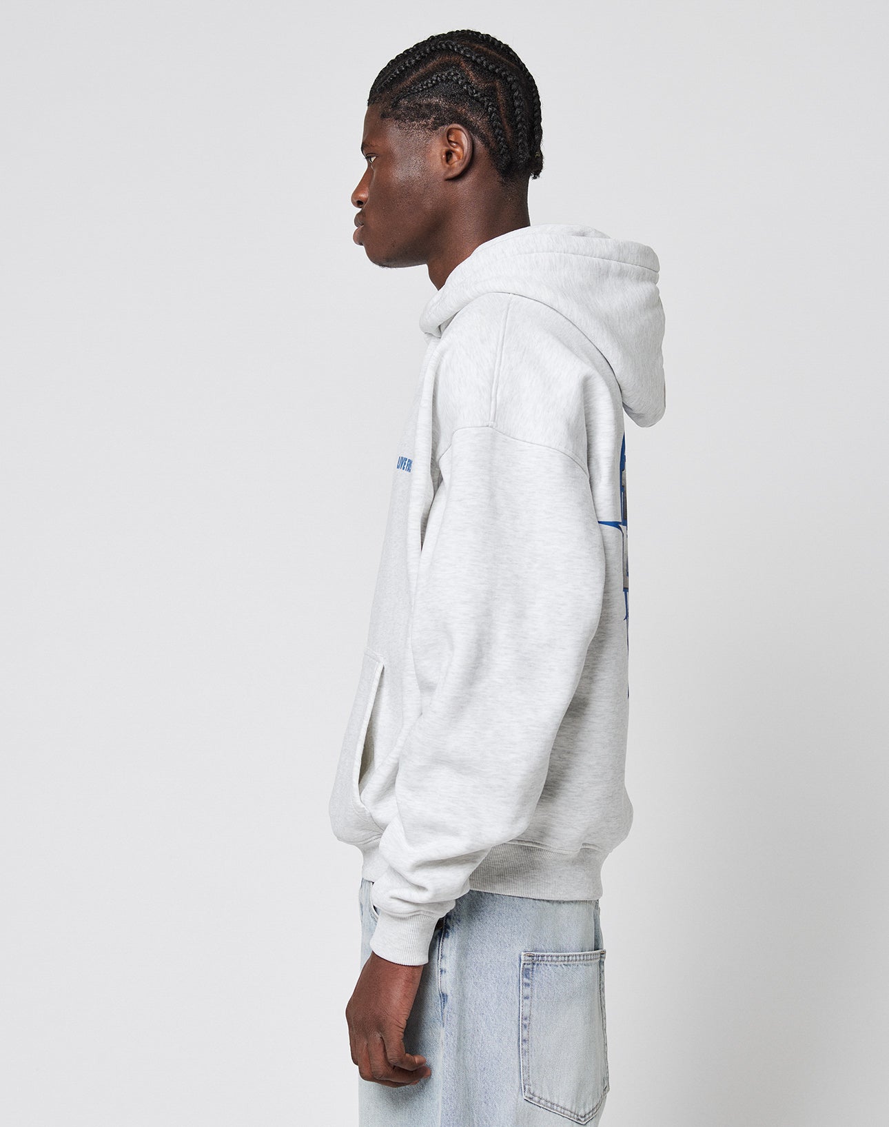 A young man with braided hair stands in profile, wearing an oversize fit light gray LFDY Globe Hooded sweatshirt by LIVE FAST DIE YOUNG, made from a Baumwollemix with Polyester, paired with light blue jeans against a white background.