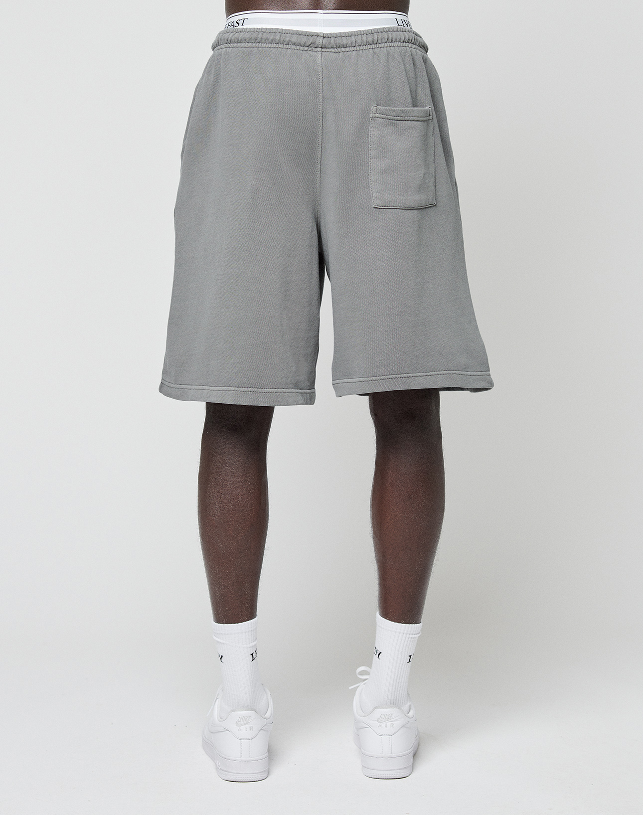 A person faces forward wearing LIVE FAST DIE YOUNG Basic Shorts in gray with a large square pocket on the right leg, paired with white socks and sneakers against a plain white background.