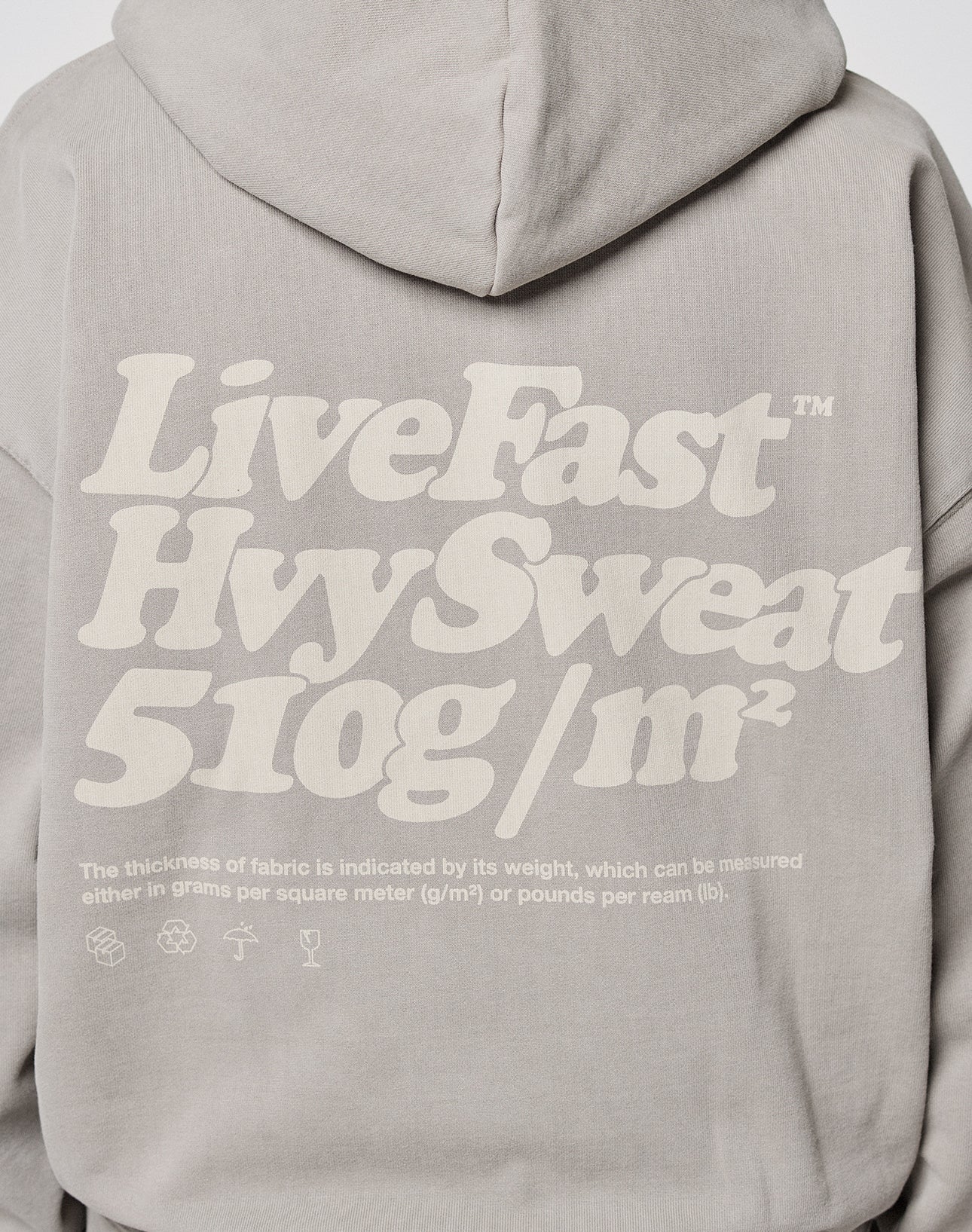 A person with a shaved head, wearing the LIVE FAST DIE YOUNG Basic 510 Hooded Zip in Oversize Fit with bold white “Live Fast Hvy Sweat 510g/m²” text on the back, stands facing away from the camera, paired with light blue jeans.| model