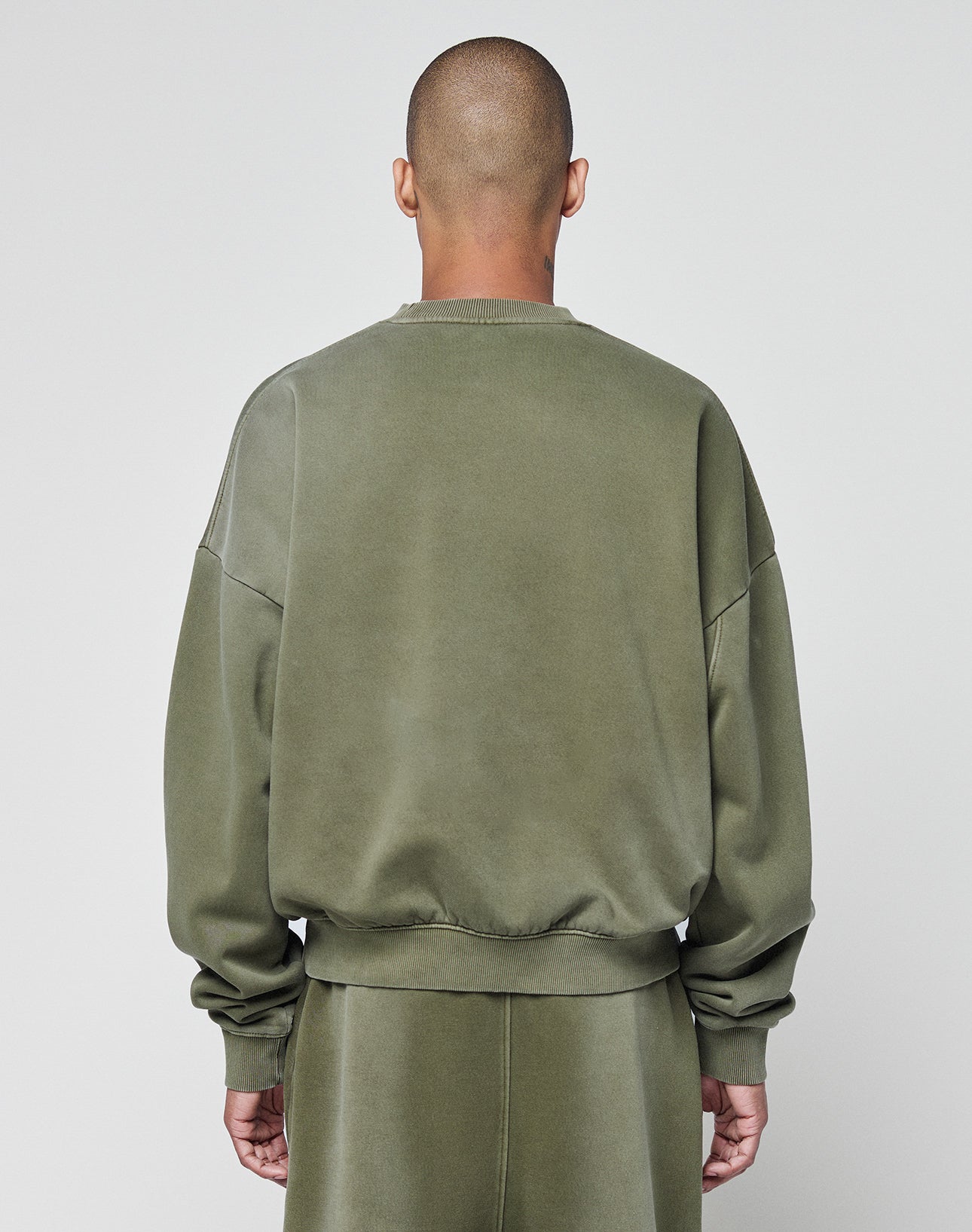 A person with a shaved head stands with their back to the camera, wearing an olive green LIVE FAST DIE YOUNG Basic Chest Sweater and matching faded pants, against a plain light gray background.