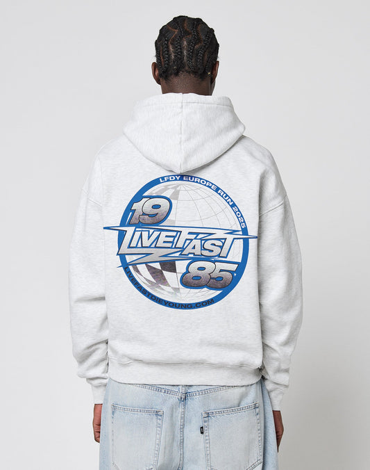 A person with braided hair is wearing the LFDY Globe Hooded by LIVE FAST DIE YOUNG—an oversize light gray hoodie. The back has a bold blue and white 19 LVETASY 85 and LFDY Europe Run 2023 graphic. The person faces away from the camera.