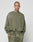 A person with a shaved head and face tattoos wears the LIVE FAST DIE YOUNG Basic Chest Sweater in oversized olive green with matching sweatpants featuring a handcrafted fade effect, standing against a plain light gray background.