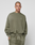 A person with short hair and visible face and neck tattoos wears the LIVE FAST DIE YOUNG Basic Chest Sweater in oversized olive green with a unique handgefertigter Fade-Effekt, standing against a plain light background.