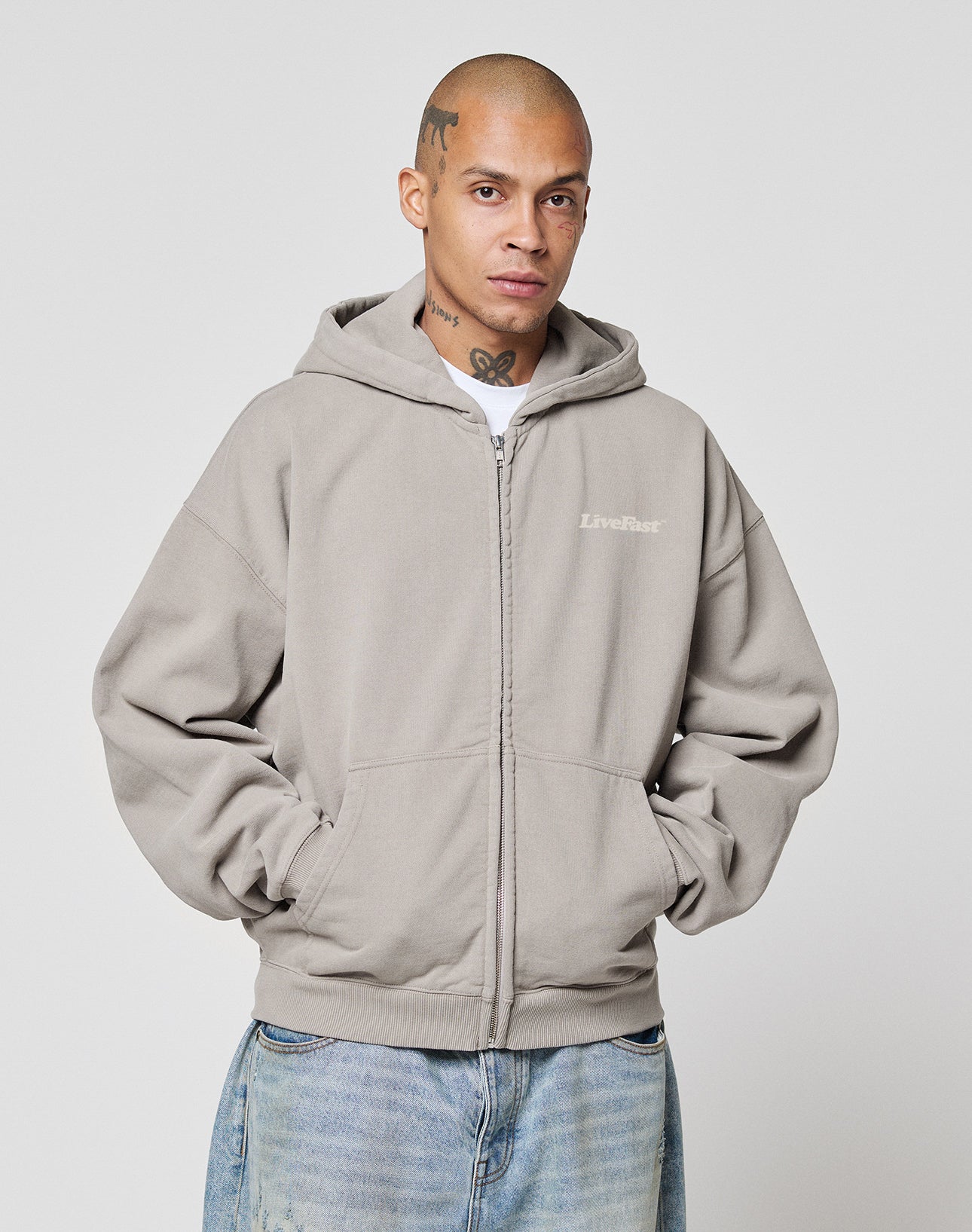 A person with a shaved head and tattoos wears the LIVE FAST DIE YOUNG Basic 510 Hooded Zip in Oversize Fit with “LiveFast” chest text, paired with light blue vintage-wash jeans, standing hands-in-pockets against a plain white background.