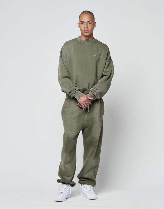 A person stands against a plain light gray background wearing the LIVE FAST DIE YOUNG Basic Chest Sweater in an oversize fit olive green, paired with matching sweatpants and white athletic shoes, posing with hands clasped in front.
