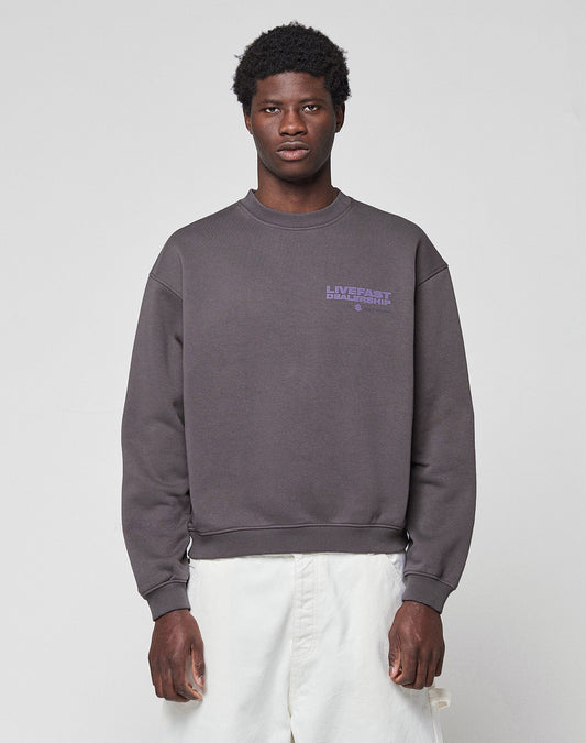 A man stands facing forward against a light gray background, wearing the LIVE FAST DIE YOUNG LFDY Network Sweater in an oversized dark gray fit with a small purple chest graphic, paired with white pants.
