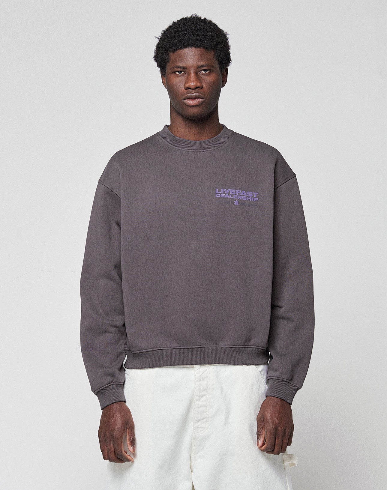 A man stands facing forward against a light gray background, wearing the LIVE FAST DIE YOUNG LFDY Network Sweater in an oversized dark gray fit with a small purple chest graphic, paired with white pants.