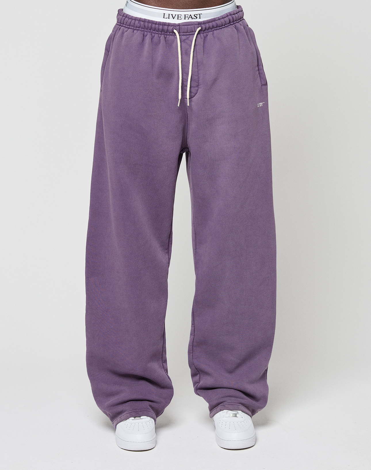A person stands wearing LIVE FAST DIE YOUNG’s Basic Wide Jogger in purple, featuring wide fit, white drawstrings, an elastic waistband with "LIVE FAST" text, and white sneakers. Background is plain and light-colored.