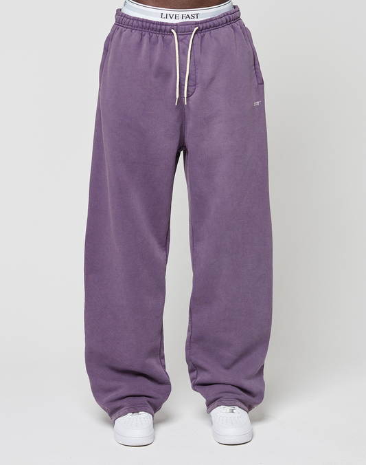 A person stands wearing LIVE FAST DIE YOUNG’s Basic Wide Jogger in purple, featuring wide fit, white drawstrings, an elastic waistband with "LIVE FAST" text, and white sneakers. Background is plain and light-colored.