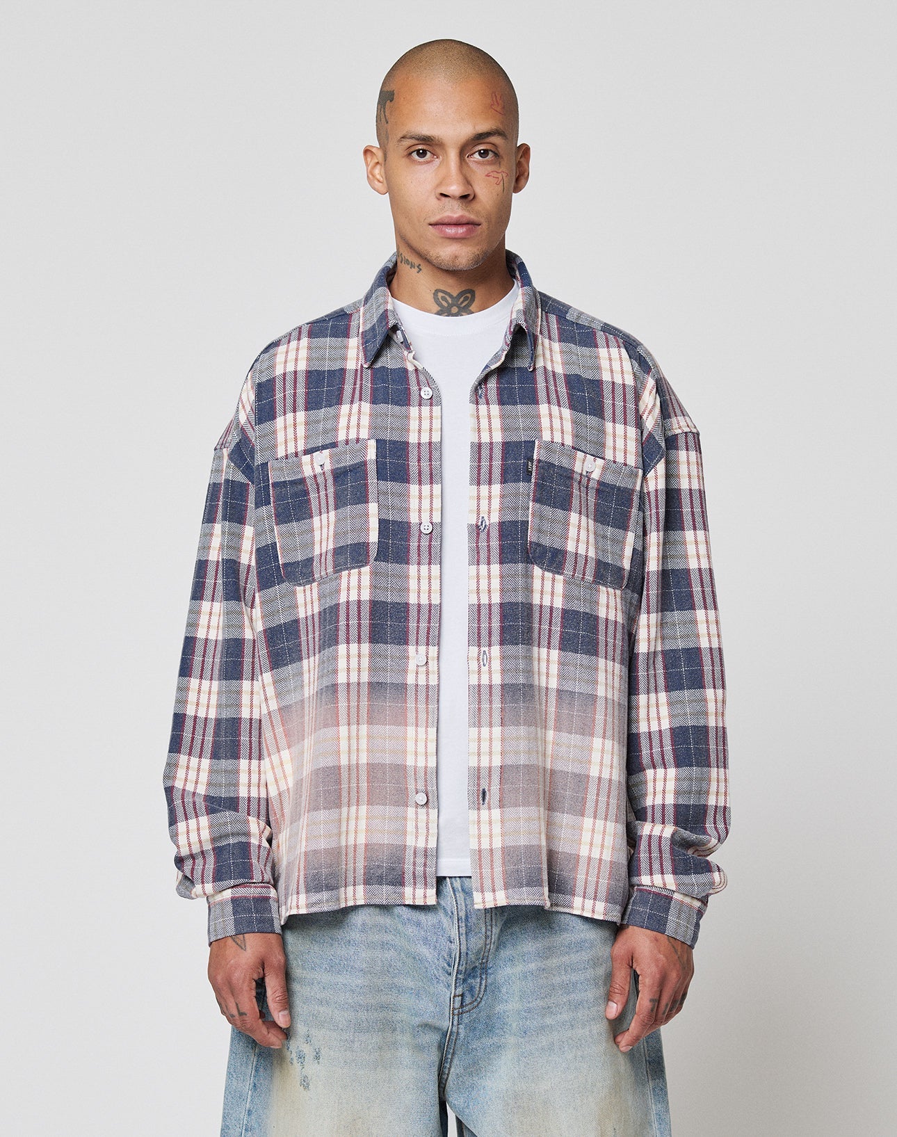 A person stands facing forward against a plain light gray background, wearing the LIVE FAST DIE YOUNG Vintage Flannel—a blue, white, and red plaid shirt with Vintage Waschung and Zweiteiliger Hemdkragen—over a white t-shirt and light blue jeans. | model