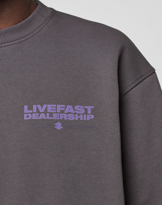 A close-up of a person wearing the LIVE FAST DIE YOUNG LFDY Network Sweater in oversize fit dark grey cotton, featuring "LIVEFAST DEALERSHIP" in light purple on the chest. Only part of their shoulder, neck, and sweater is visible.