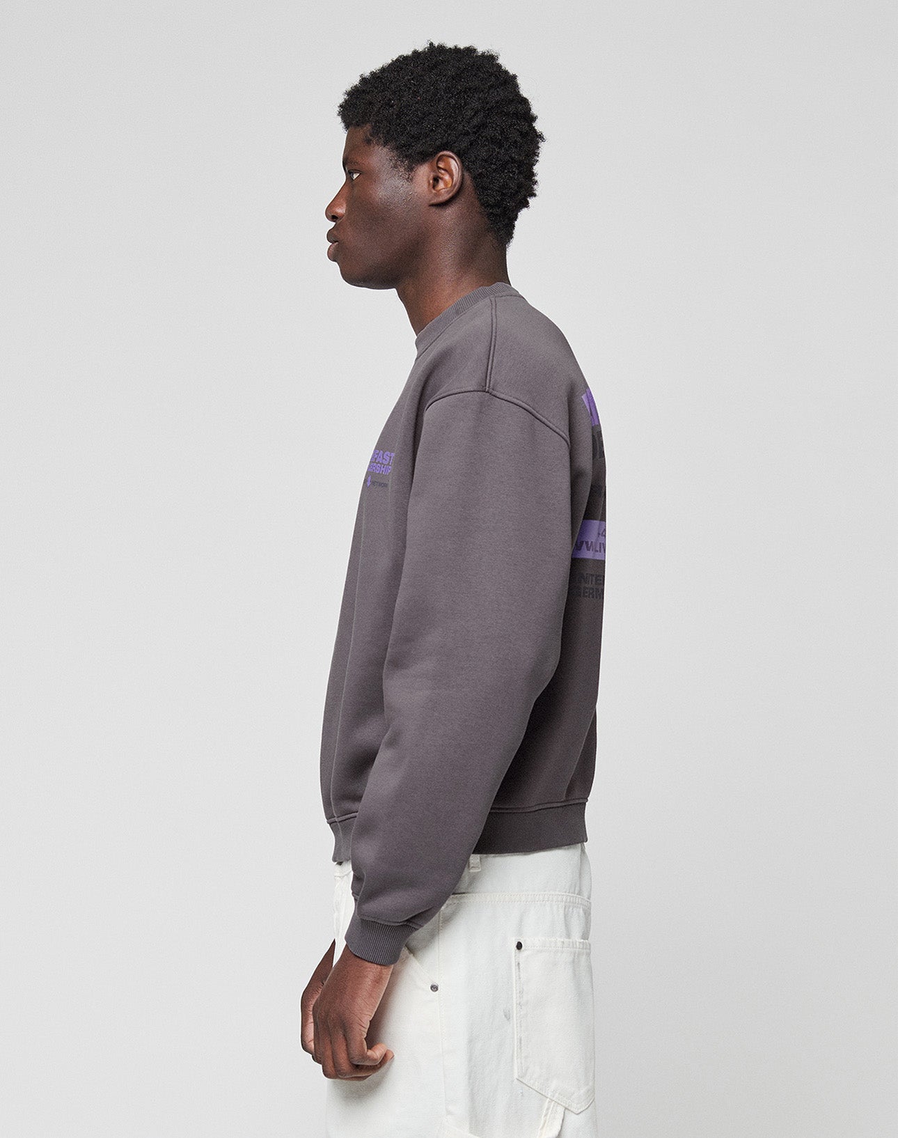 A person in profile faces left, wearing the LIVE FAST DIE YOUNG LFDY Network Sweater—an oversized dark grey cotton sweatshirt with purple front and back prints—paired with light pants, set against a plain light grey background.
