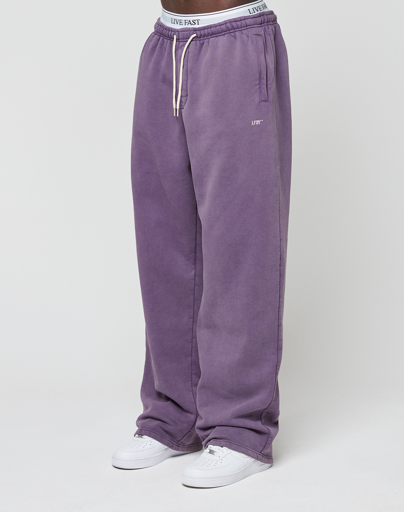 A person wears LIVE FAST DIE YOUNG Basic Wide Joggers in loose purple with a white drawstring and "LIVE FAST" elastic waistband, paired with white sneakers against a plain white background. | model