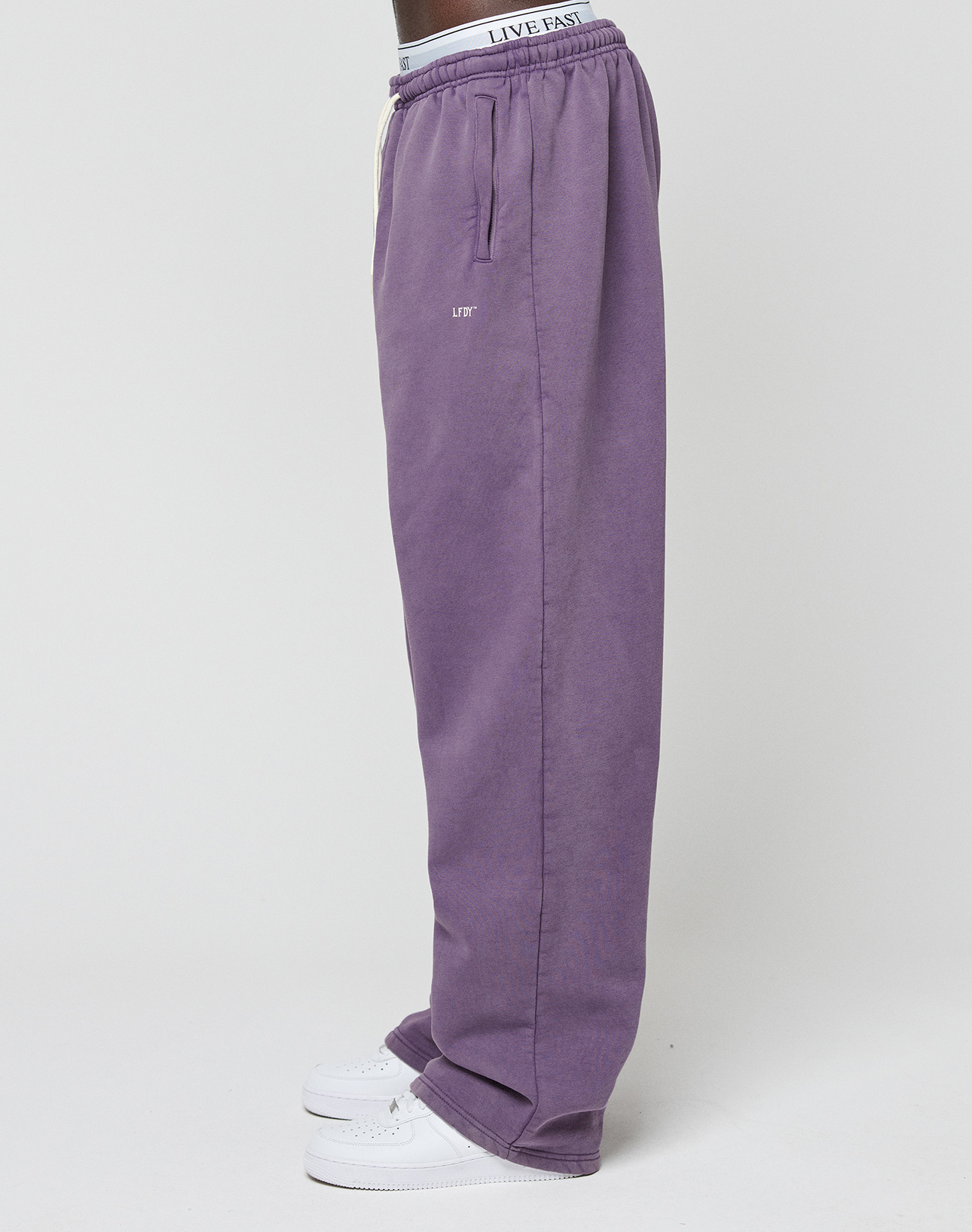 A person stands sideways against a plain light background wearing LIVE FAST DIE YOUNG Basic Wide Jogger in purple, white sneakers, and underwear featuring a visible elastikbund waistband with the text LIVE EAST.