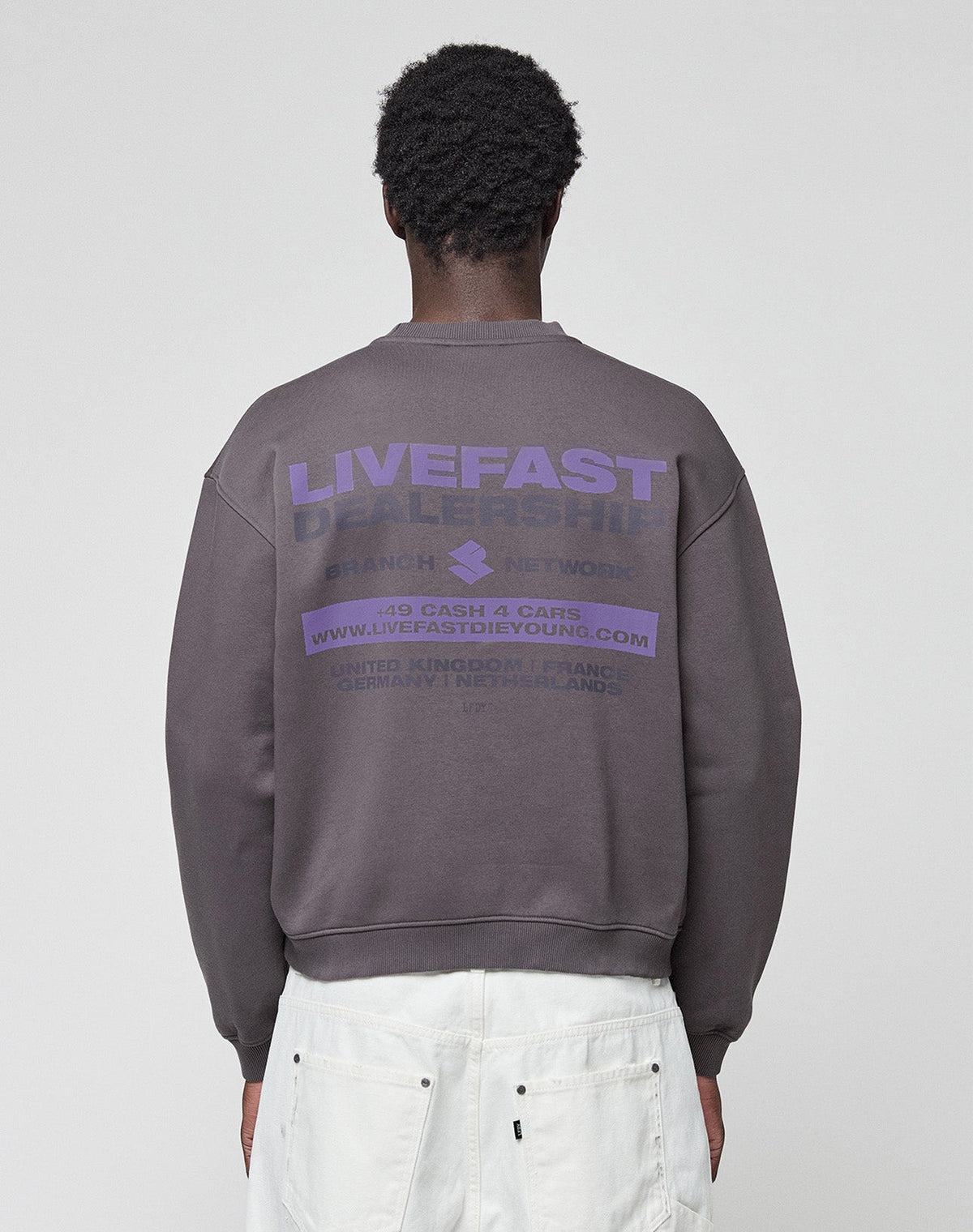 A person faces away, wearing the LFDY Network Sweater by LIVE FAST DIE YOUNG—an oversize dark gray cotton sweatshirt with purple "LIVEFAST DEALERSHIP" front and back prints—paired with white pants, set against a plain light background. | model