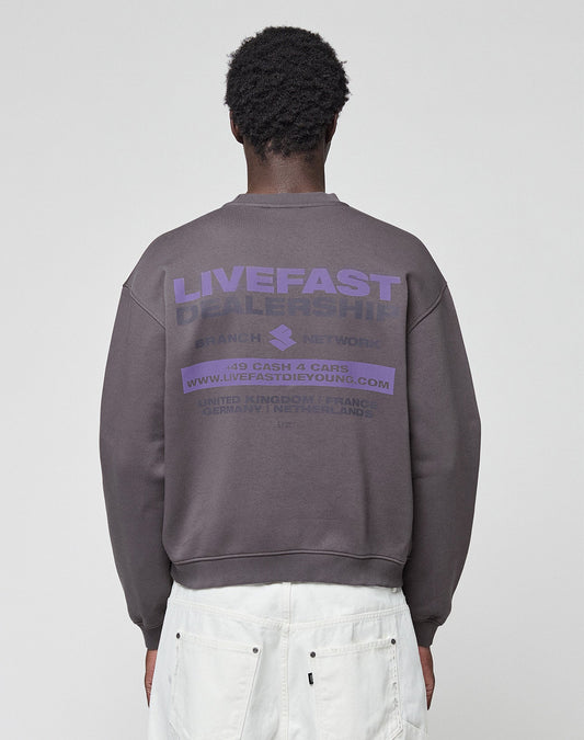 A person faces away, wearing the LFDY Network Sweater by LIVE FAST DIE YOUNG—an oversize dark gray cotton sweatshirt with purple "LIVEFAST DEALERSHIP" front and back prints—paired with white pants, set against a plain light background. | model